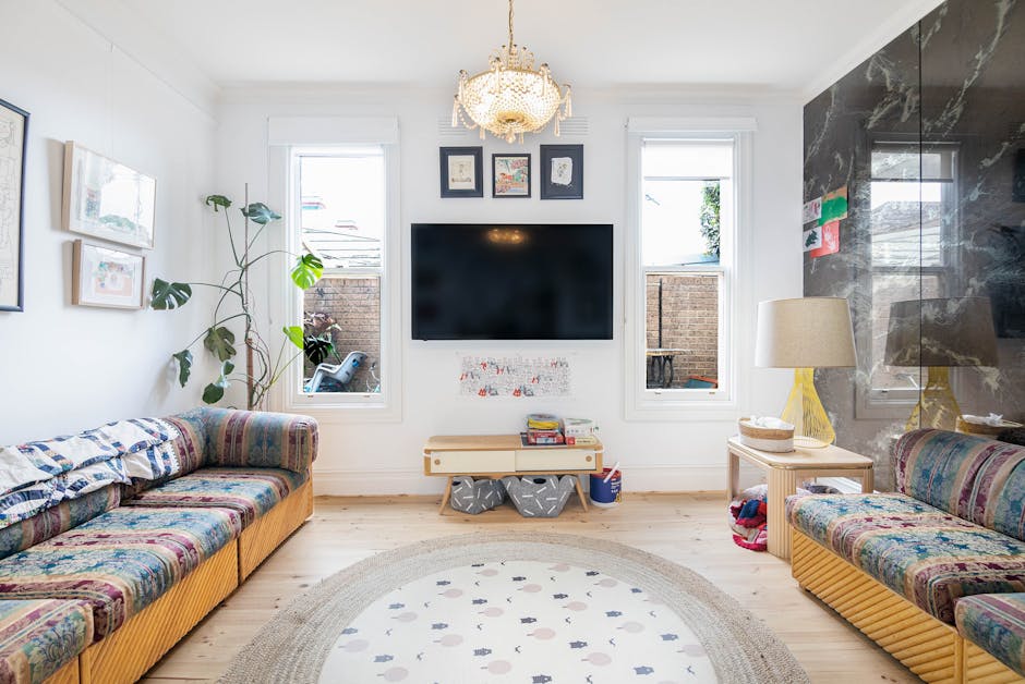 A cozy living room with a bright, airy atmosphere illuminated by natural light coming through two large windows with simple white frames. The room features two vintage-style sofas with colorful, patte