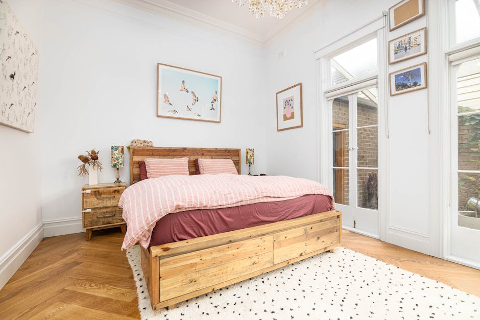 A cozy living room with a bright, airy atmosphere illuminated by natural light coming through two large windows with simple white frames. The room features two vintage-style sofas with colorful, patte