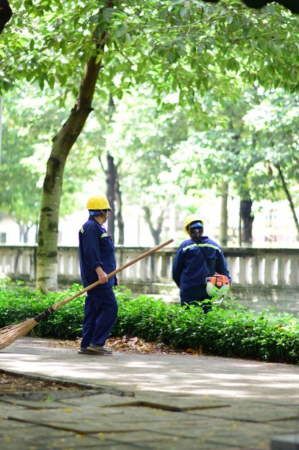 A young woman and a male gardener are walking along a narrow pathway between lush, well-maintained garden beds filled with green, foliage plants. The woman is holding a blue spray bottle and wearing g